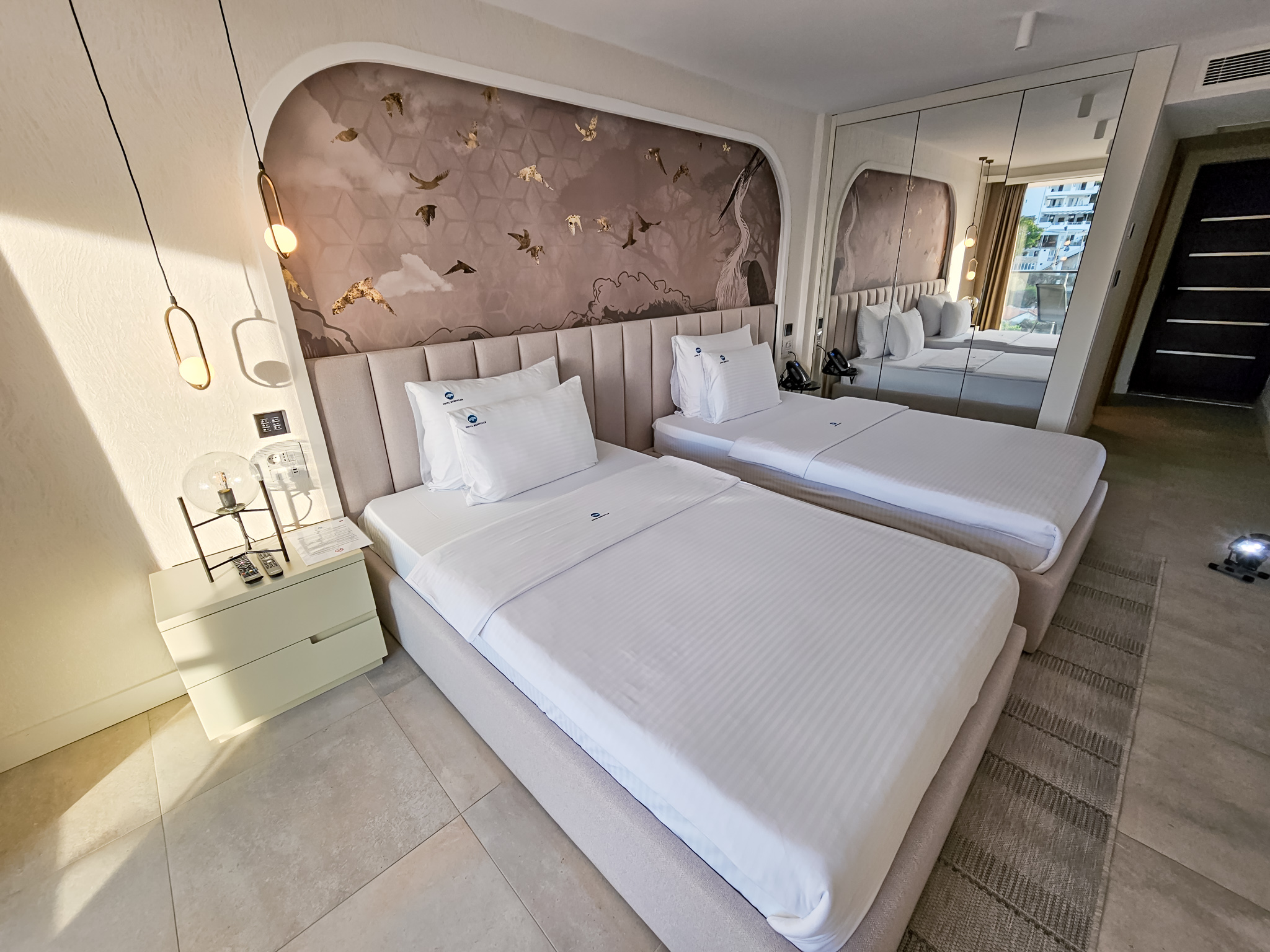 Moderna hotelska soba sa dva kreveta, elegantnom taupe glavnom naslonjaču sa zlatnim pticama i ogledalom