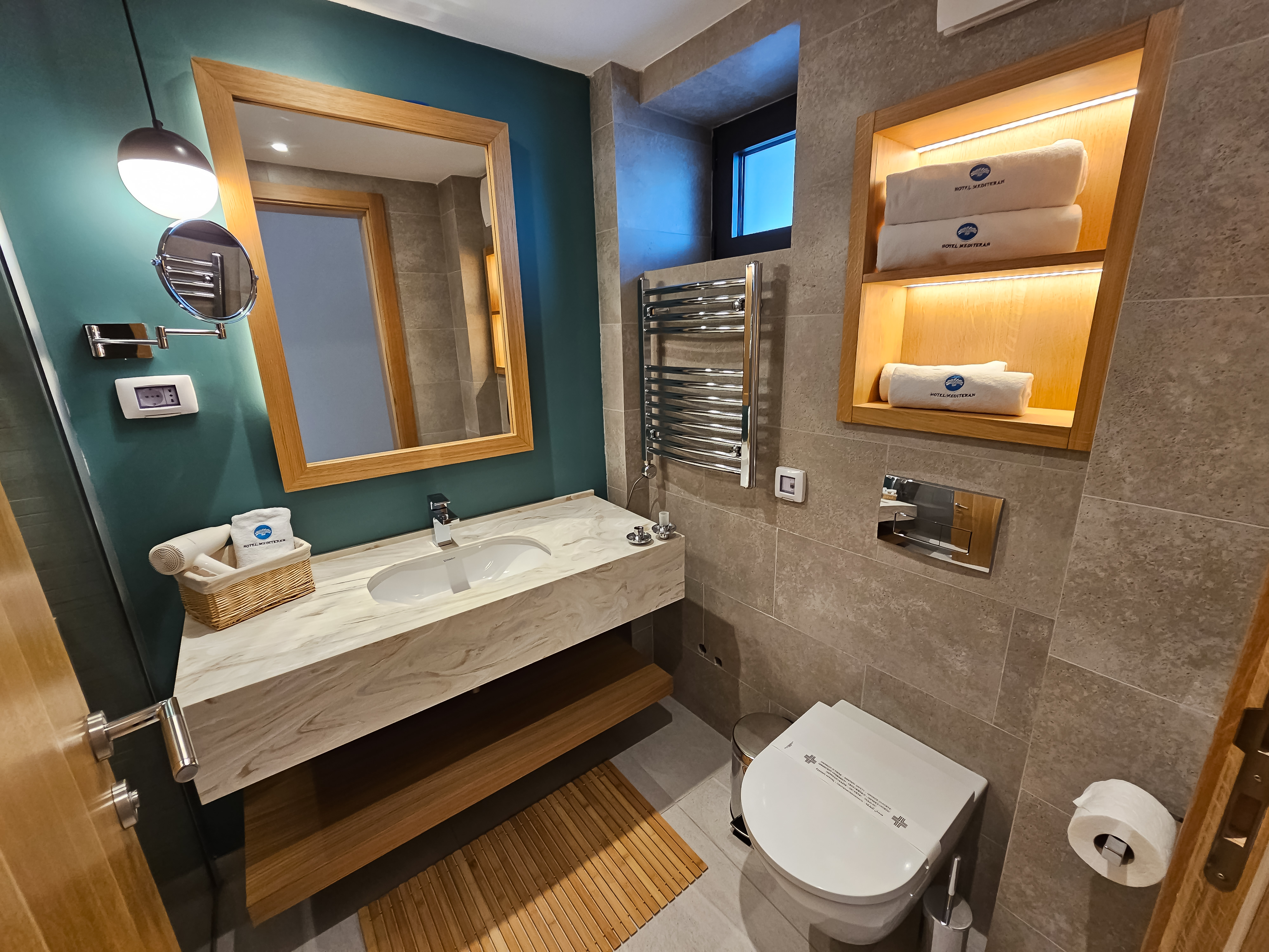 Moderan hotelski kupatilo sa tirkiznom akcentnom zidnom, mramornim umivaorom i osvetljenim nicama sa peškiri