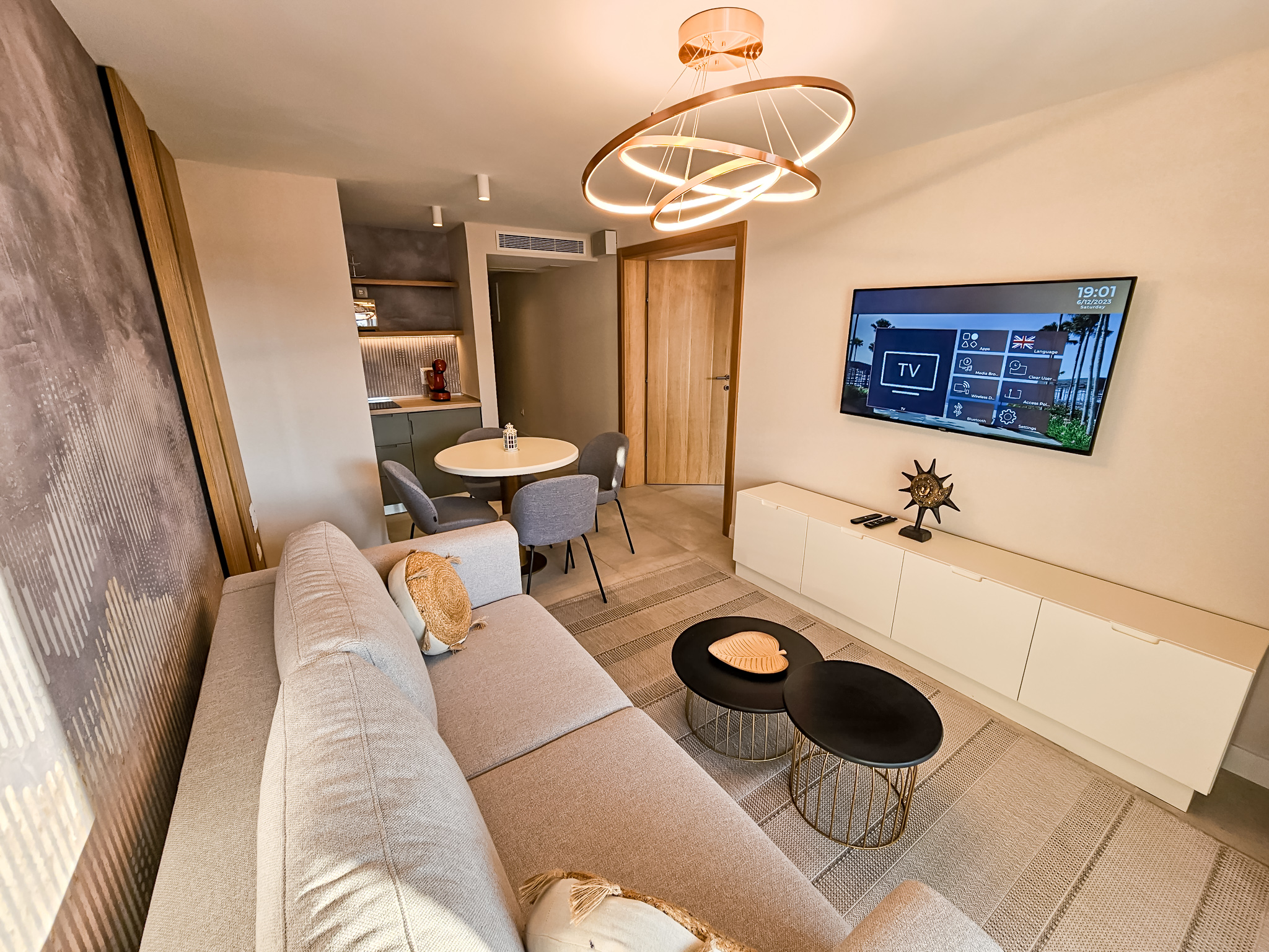 Moderna hotelska suite sa sivom sofom, kuhinjom i blagavaonom, osvetljena zlatnom lustre